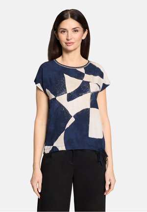 Kobieta w granatowo-białej bluzce z krótkim rękawem i abstrakcyjnym wzorem, z okrągłym dekoltem, ubrana w czarne spodnie, stoi na tle jednolitego tła.
