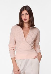 Helles rosa Wickelsweatshirt mit gerippter Struktur, langen Ärmeln, V-Ausschnitt und dezentem Kabelstrickmuster, kombiniert mit cremefarbenen Hosen.