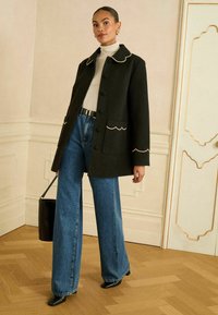 Zwarte mantel van wolmix met geschulpte halslijn en zakaccenten, gecombineerd met wijde blauwe denim jeans en zwarte schoenen met puntige neus.