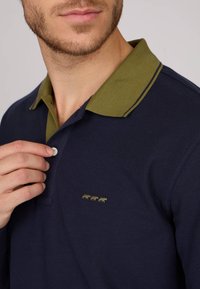 Polo shirt bleu marine avec un col vert et des bordures jaunes, arborant un motif brodé à trois figures sur la poitrine. Détail du col à boutons visible.