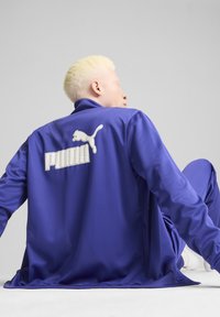 Puma dzseki élénk lila színben, sima textúrával, magas gallérral, és egy nagy fehér logóval a hátsó részen. A modell egy semleges háttér előtt ül.