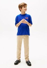 Blaues Baumwoll-T-Shirt mit einem kleinen roten und weißen Patch, beigen Hosen und schwarzen Slippern. Das Shirt hat eine lockere Passform und kurze Ärmel.