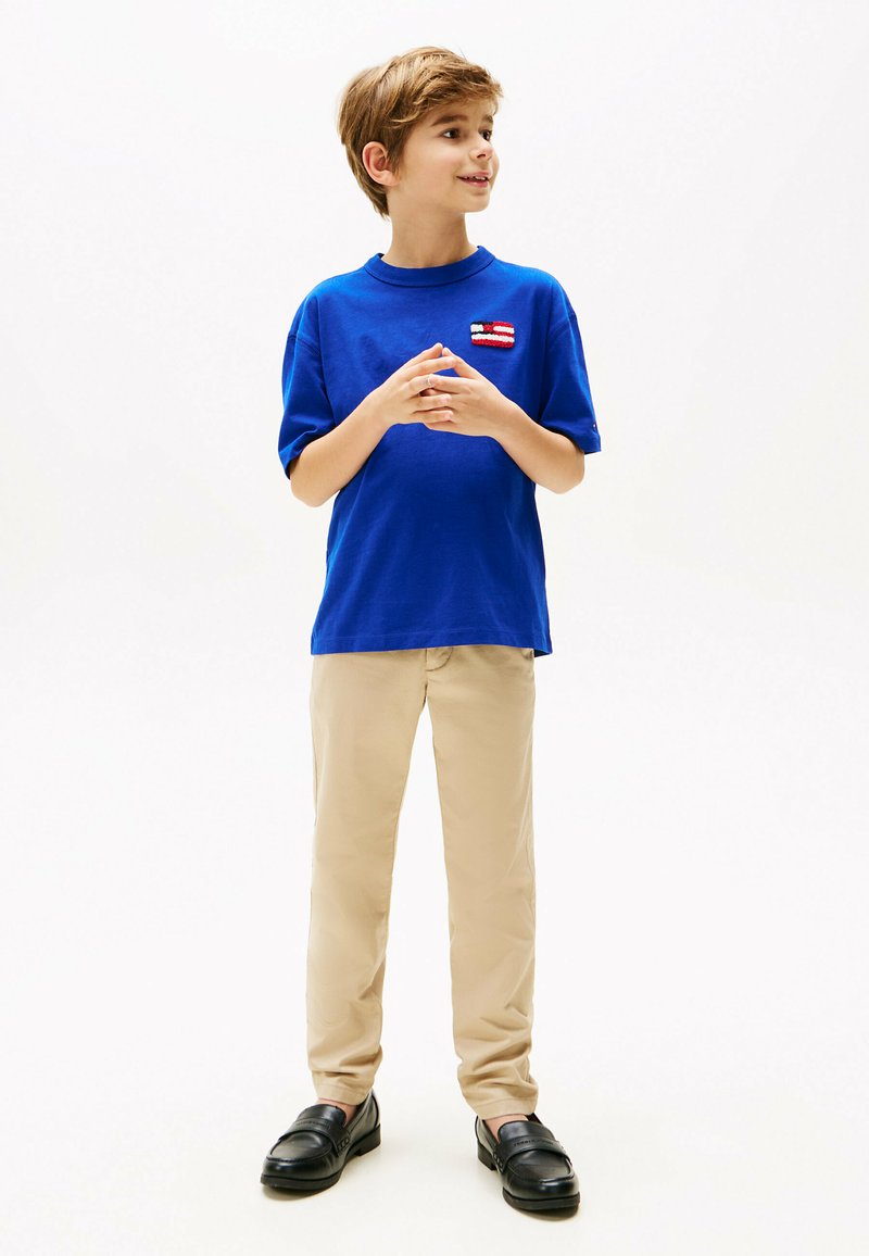 Blå bomulds t-shirt med et lille rød-hvidt mærke, beige bukser og sorte loafers. T-shirten har en afslappet pasform og korte ærmer.