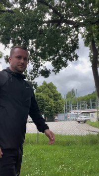 Black nylon jacket with a hood, standing figure in front of green grass and trees, gray clouds in the sky, partial view of a parking lot.