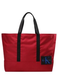 Borsa tote in tessuto rosso con manici neri, forma rettangolare, con un'etichetta logo blu e nera con la scritta "Calvin Klein Jeans." Texture liscia.