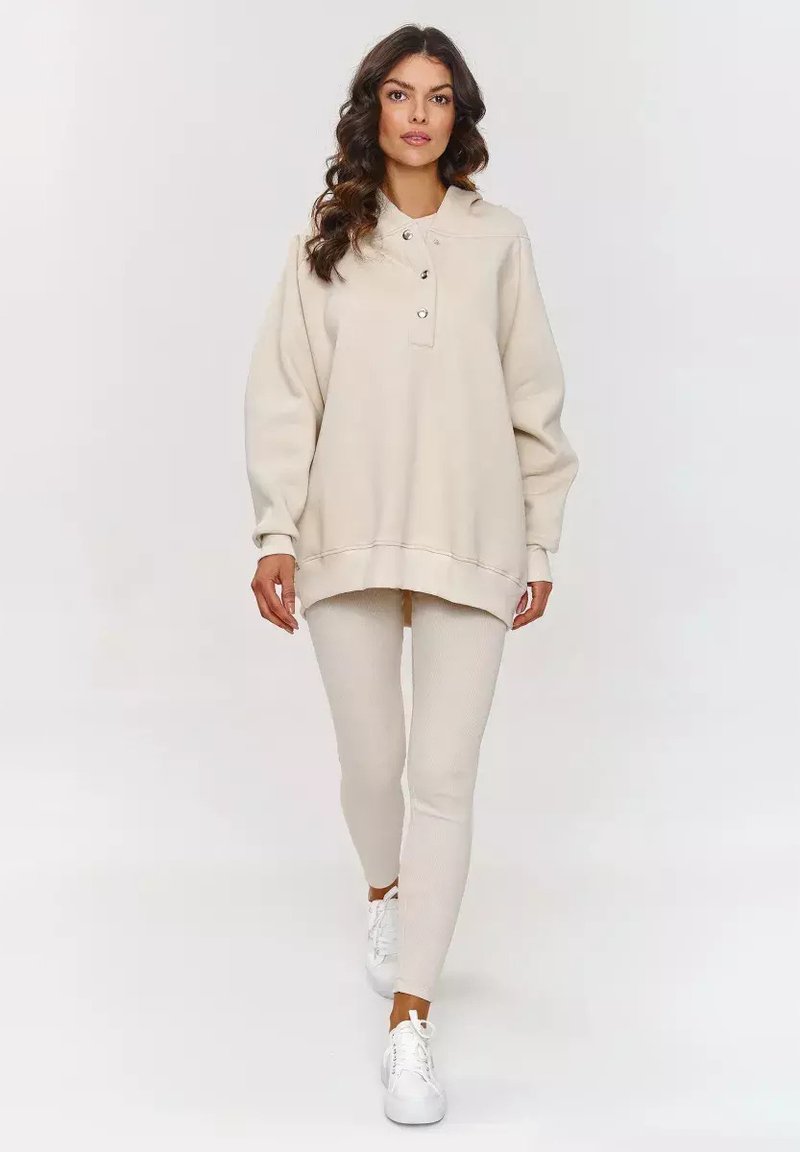 Beżowy, oversize'owy sweatshirt z kapturem, czteroma guzikami oraz ściągaczami, w połączeniu z teksturowanymi, beżowymi legginsami i białymi sneakersami.