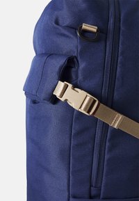 Marineblå ryggsekk med teksturert stoff, en sidelomme med glidelås og en lysebrun spenne for sikker lukking.