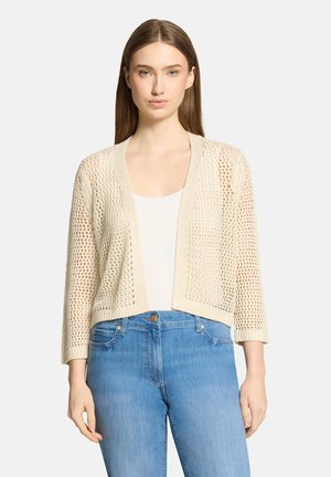 Jeune femme portant un cardigan crème à mailles ajourées avec des manches trois-quarts par-dessus un haut blanc et un jean bleu taille haute.