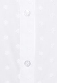 Blusa di cotone bianca con un motivo testurizzato di piccoli fiori. Presenta una chiusura con bottoni frontali a pressione, dotata di bottoni rotondi e trasparenti.