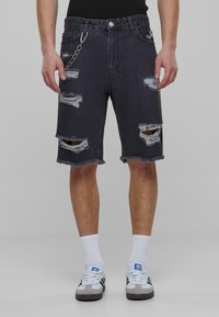 Pantalones cortos de mezclilla negros con desgastes, dobladillo deshilachado y detalle de cadena en el lazo del cinturón, combinados con calcetines blancos y zapatillas grises.