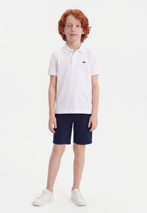 Rødhåret dreng står iført en hvid poloshirt, navyblå shorts og hvide sneakers mod en ensfarvet hvid baggrund.