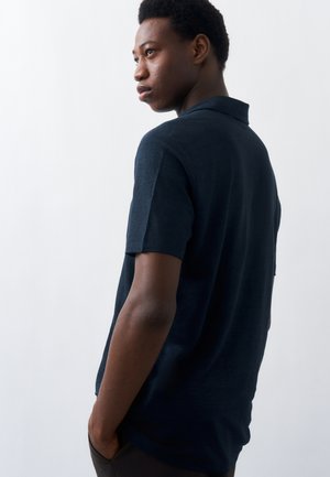 Mladi moški, oblečen v temno modro polo majico s kratkimi rokavi, stoji z rokami v žepih in gleda v stran na enostavni svetli ozadju.