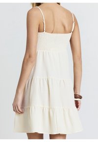 Robe à dos froncé de couleur crème avec fines bretelles, design à volants et tissu texturé. Caractérisée par un corsage ajusté et une jupe fluide.