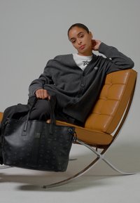 Eine Frau in einer grauen Strickjacke und dunklen Hosen sitzt auf einem hellbraunen Ledersessel und hält eine große schwarze Tragetasche mit Muster.