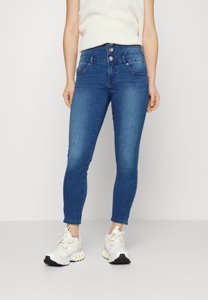 Slim-fit blue jeans with three buttons at the waist worn with a white knit sleeveless top and white chunky sneakers.