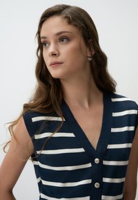 Marinblauwe mouwloze top met witte verticale strepen, V-halsontwerp en drie zilveren knopen langs de voorkant, gladde breistek.