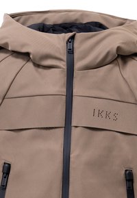 Chaqueta beige con capucha, cremalleras negras y logo de IKKS en el pecho, con dos bolsillos con cremallera y forro interior acolchado.