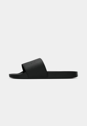 Sorte sandaler med bred, teksturert rem og flat såle, vist fra en sideprofil mot en ensfarget hvit bakgrunn.