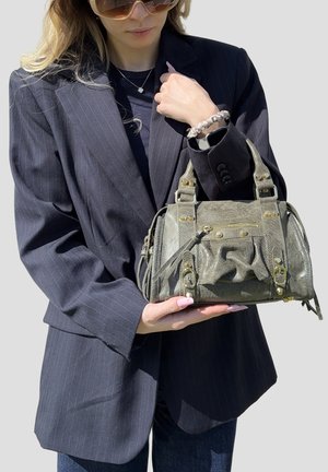 Donna con occhiali da sole e blazer a righe tiene una borsa verde oliva testurizzata con dettagli dorati davanti a uno sfondo grigio.