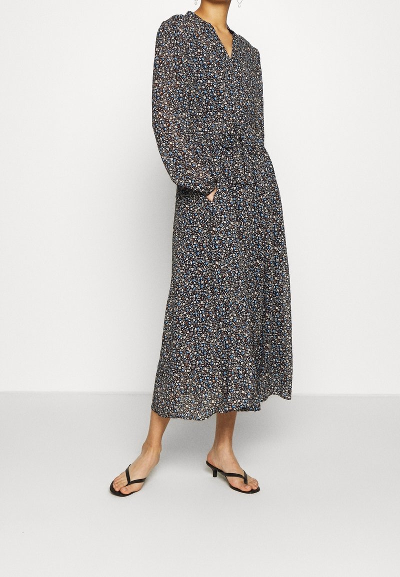 Femme portant une robe midi noire à manches longues avec de petites fleurs bleues et blanches et des sandales noires à talons bas, debout avec une main dans la poche.