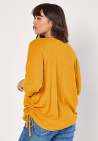 Senapsgul överdimensionerad topp, ribbad textur, långa ärmar, rynkat detaljer vid sidorna, utställd nederkant, matchad med blå jeans.