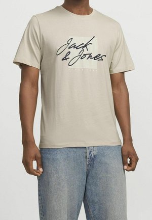 Homme portant un t-shirt beige à manches courtes avec le texte "Jack & Jones" et un jean denim bleu clair, debout devant un fond uni.