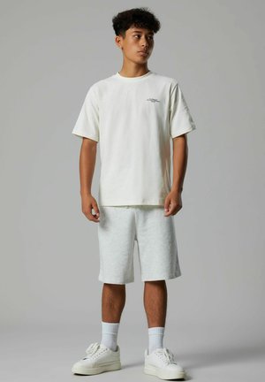 Giovane uomo in piedi, indossa una t-shirt bianca oversize, pantaloncini grigio chiaro, calzini bianchi e scarpe da ginnastica bianche su uno sfondo grigio uniforme.