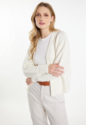Weißer Strickcardigan mit lockerer Passform, gerippten Bündchen und offener Front, kombiniert mit einem leichten Oberteil und beigen, taillierten Hosen.