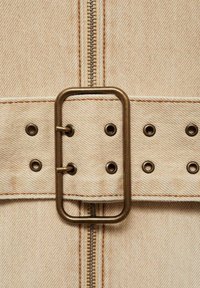 Bež denim odjeća koja uključuje brončanu pravokutnu kopču za pojas, vidljivi patentni zatvarač i detalje šivanja s metalnim rupicama.