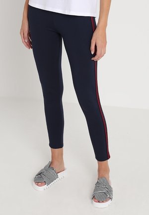 Marineblauwe leggings met rode zijstrepen, gemaakt van rekbaar materiaal. Gepaard met zwart-wit gestreepte instapschoenen met een gestructureerde zool.