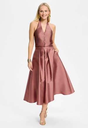 Femme blonde portant un haut halter en satin rose sans manches avec des boutons dorés et une jupe midi évasée assortie, des boucles d'oreilles dorées, un bracelet et des talons.