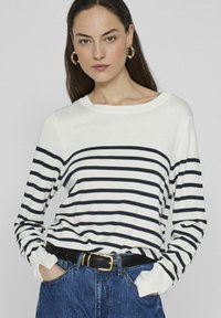 Camiseta blanca de manga larga con rayas horizontales marino, cuello redondo y puños acanalados; combinada con jeans azul de cintura alta y un cinturón negro.