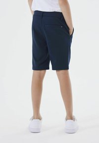 Pantaloni corti blu navy con una texture liscia e una tasca posteriore; indossati con sneakers bianche; mostrati da dietro, su uno sfondo bianco uniforme.