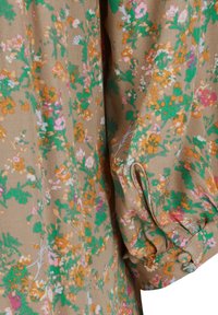 Kukallinen kangas maanläheisessä ruskeassa, jossa on vihreitä, vaaleanpunaisia ja oransseja kukkia. Pehmeä rakenne, jossa on kerätty hihojen yksityiskohta ja napin koriste.