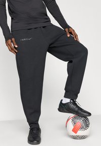 Calças de moletão pretas da Nike com "PARIS" em cinza, corte descontraído, parte de um traje desportivo. Um sapato preto, em pé sobre uma bola de futebol com padrão.