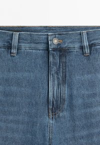 Close-up of blue denim jeans waistband with belt loops, button closure, and visible zipper under the flap.