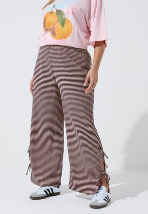 Person iført lyserød oversized T-shirt med ferskenmotiv og løse brune bukser med striber og knyttede sideslids, kombineret med hvide og sorte sneakers.