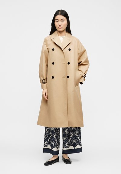Γυναίκα με μακριά σκούρα μαλλιά φοράει ένα μπεζ διπλόπατο παλτό τύπου trench, παντελόνι με φαρδιά μπατζάκια και σχέδιο, και μαύρα μπαλαρίνες, στέκεται μπροστά σε λευκό φόντο.