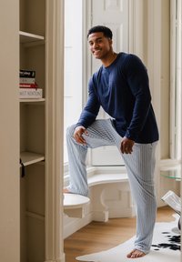 Marineblauw shirt met lange mouwen en een gedrukt logo, gecombineerd met lichtblauwe pyjamabroek met strepen. Houten vloer en witte planken zijn zichtbaar.