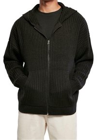 Svart ribbad zip-up hoodie med stor huva, framfickor och texturerat tyg. Bärs över en svart skjorta med krämfärgade byxor.