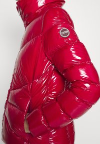 Skinande röd pufferjacka med hög krage, quiltat mönster och en cirkulär logotyppatch på vänster ärm.