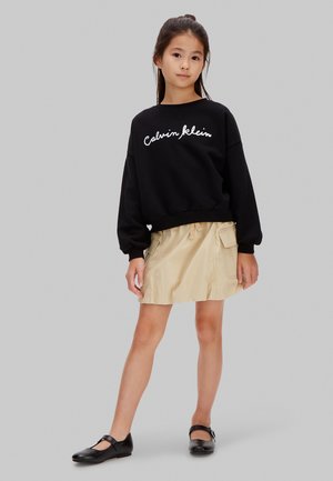 Jeune fille debout portant un sweat-shirt noir Calvin Klein, une jupe beige avec poches, et des chaussures Mary Jane noires sur un fond gris.