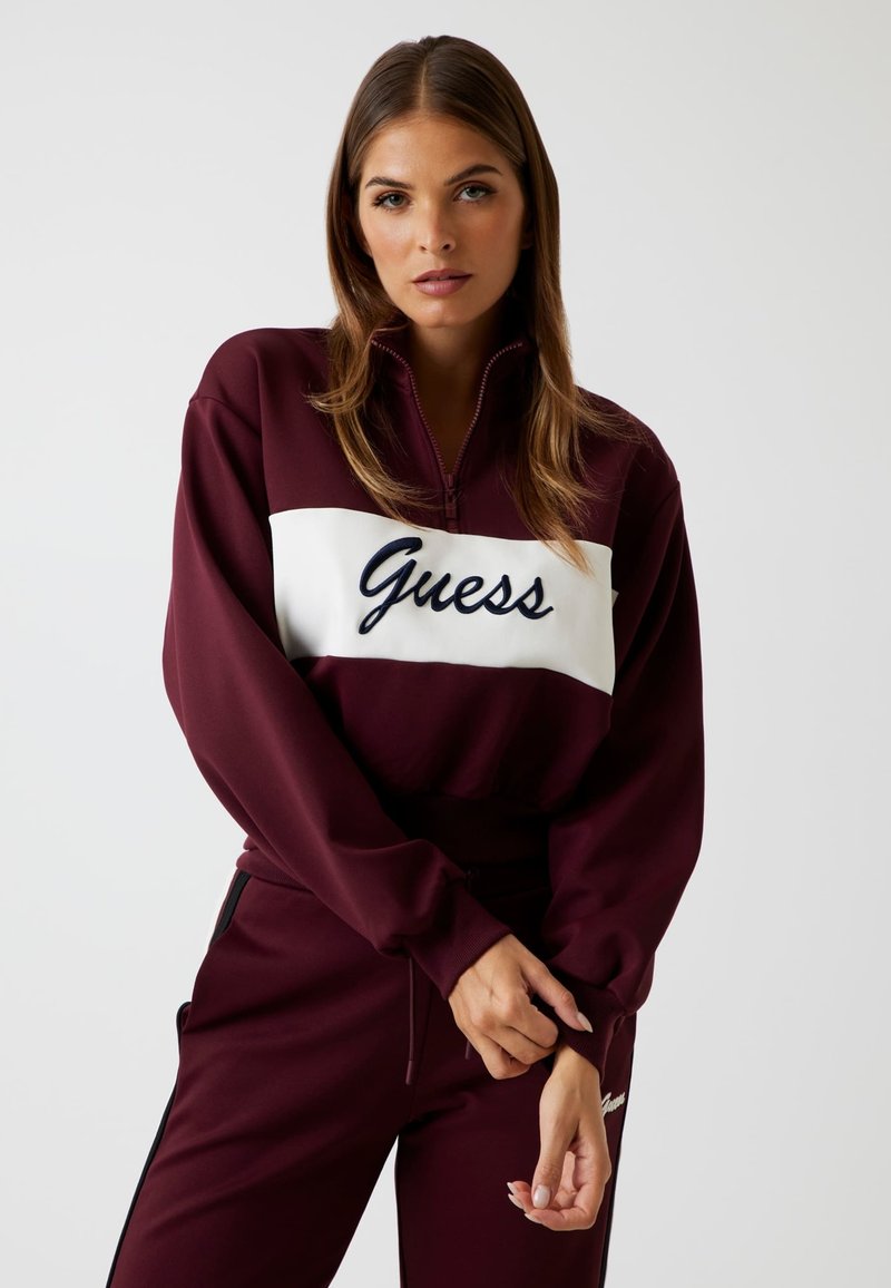 Felpa con zip color bordeaux con pannello bianco che presenta "Guess" in corsivo navy, abbinata a pantaloni coordinati con dettagli neri. Tessuto morbido, vestibilità rilassata.