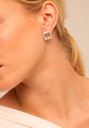 Pendientes de botón cuadrados de plata con un acabado texturizado y centro abierto, asegurados firmemente, mostrados en la oreja de una modelo.