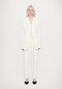 Blazer blanc ajusté avec des épaules larges, assorti à un pantalon large. Tissu texturé, fermeture à un bouton. Chaussures noires à talons pointus.