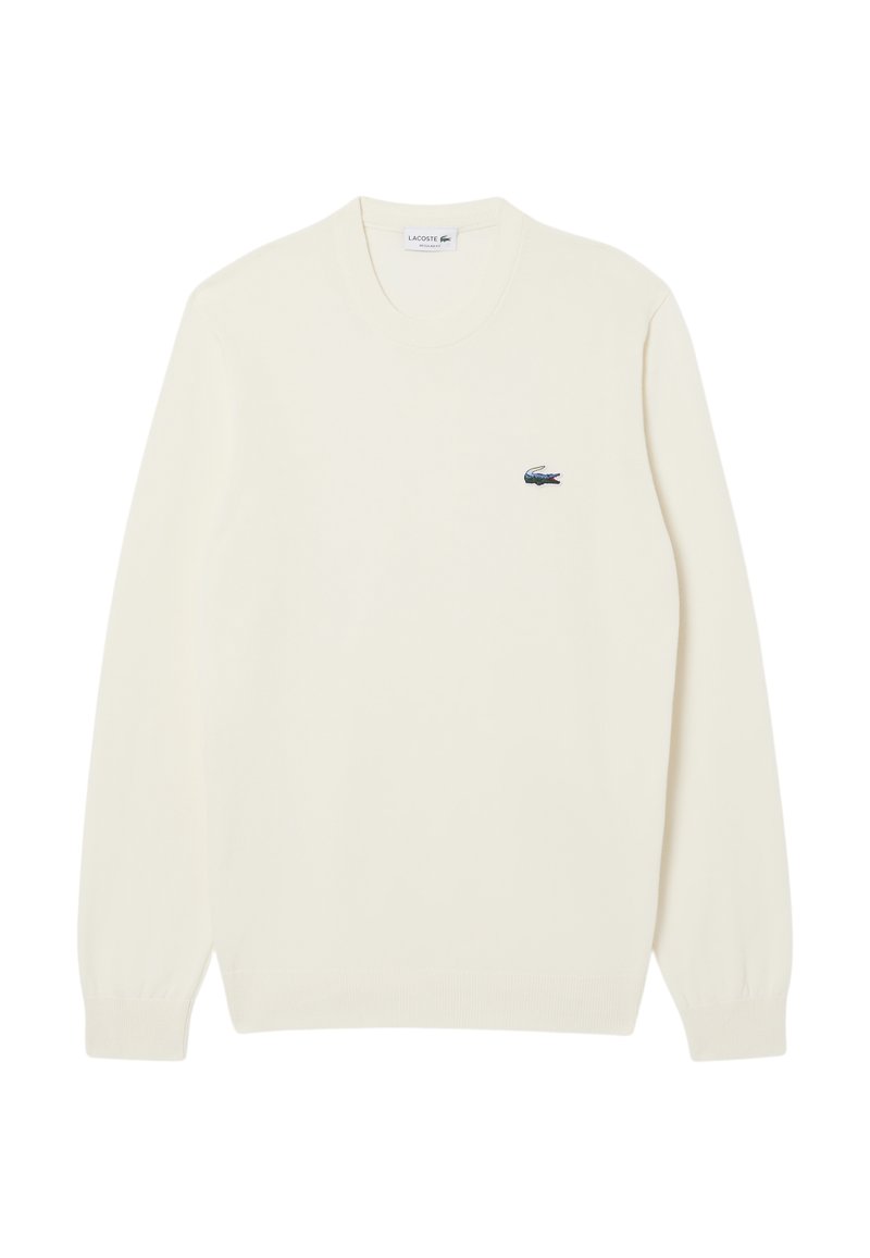 Pull crème avec un col ras du cou et des poignets côtelés. Affiche un petit logo Lacoste bleu et vert sur la poitrine gauche.