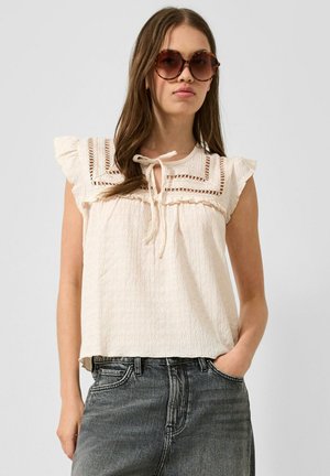 Femme portant de grandes lunettes de soleil rondes, un chemisier sans manches crème avec des détails en dentelle et un jean noir délavé taille haute, une main dans la poche.