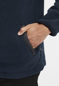 Marineblå fleece-pullover med struktureret stof, sidelomme med sort kant, justerbar snor i kanten og en afslappet pasform.