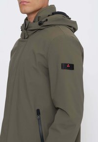 Giacca verde oliva con cappuccio, tasche laterali con cerniera e una toppa di tessuto nero con logo rosso sulla manica sinistra. Texture liscia.