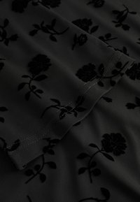 Čierna látka s textúrovaným kvetinovým vzorom. Obsahuje mäkké, sametové kvety a listy v čiernej farbe na hladkom, priehľadnom pozadí.